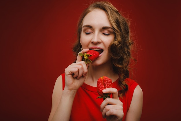 Una bella ragazza in un vestito rosso su un rosso tiene una fragola tra le mani e sorride.