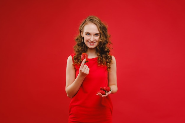 Una bella ragazza in un vestito rosso su uno sfondo rosso tiene una fragola tra le mani e sorride.