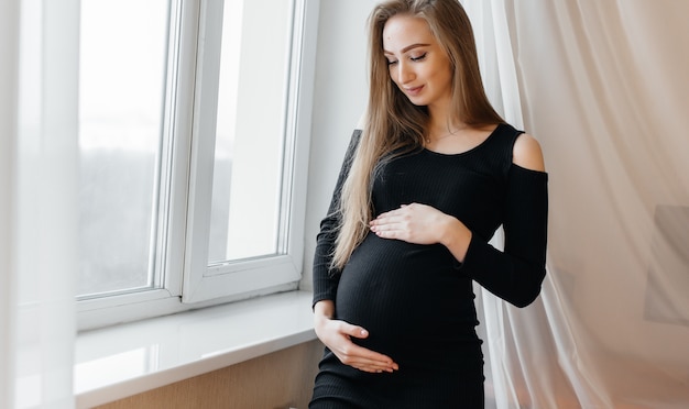 Una bella ragazza incinta è in piedi in una stanza vicino alla finestra.