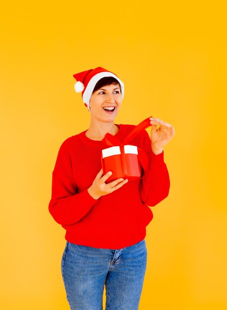 Una bella ragazza sexy con un cappello di Capodanno e un maglione rosso che tiene un regalo tra le mani. Festeggiamo il Natale o il Capodanno su uno sfondo giallo