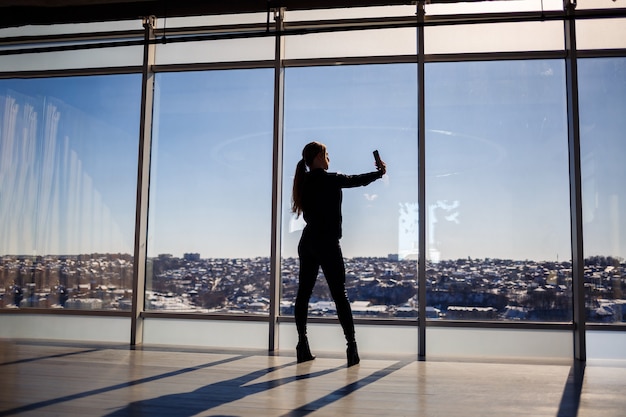 Una bella ragazza si fa un selfie con il telefono sullo sfondo delle finestre panoramiche di un grattacielo. Messa a fuoco selettiva