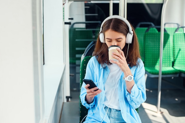 Una bella ragazza siede nel moderno tram o metropolitana della città, ascolta musica e beve il caffè.