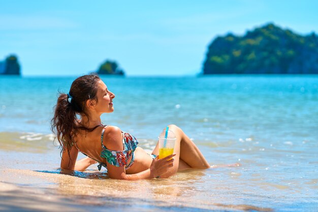 Una bella ragazza snella in costume da bagno si gode l'abbronzatura sdraiata su una spiaggia tropicale, con in mano un bicchiere di succo d'ananas.