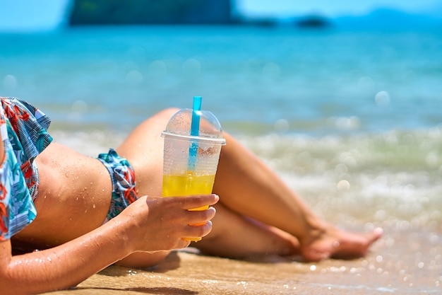 Una bella ragazza snella in costume da bagno si gode l'abbronzatura sdraiata su una spiaggia tropicale, con in mano un bicchiere di succo d'ananas.