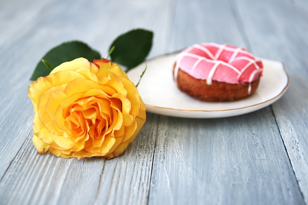 Una bella rosa gialla con un bocciolo aperto e una ciambella con glassa rosa su un piatto di ceramica bianco su un legno grigio