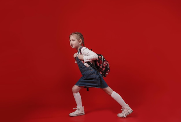 Una bella scolaretta in abito di jeans con uno zaino sulle spalle cammina su uno sfondo rosso isolato.