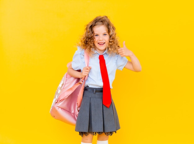 Una bella scolaretta in uniforme scolastica, con un sorriso felice. Studentessa carina con capelli biondi che tiene in mano una valigetta rosa su sfondo giallo isolato isolated