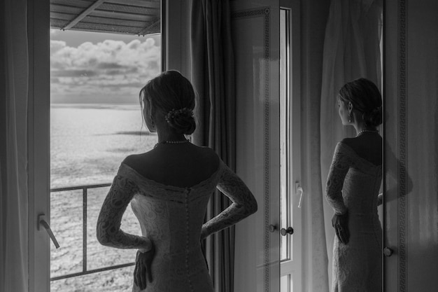 Una bella sposa snella in abito bianco guarda la scena del mare dalla sua finestra nell'hotel