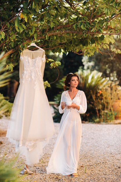 Una bella sposa sta accanto a un abito da sposa con una tazza di tè in un abito da boudoir accanto a una villa in Italia.mattina della sposa in Toscana.