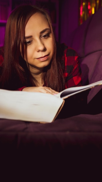 Una bella studentessa che legge un grande libro Una giovane donna premurosa sdraiata sul divano con un libro e la sera legge in una stanza buia