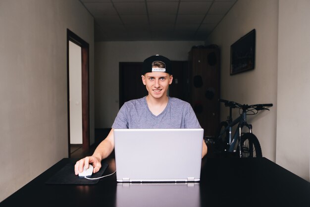 Una bella studentessa sorridente con un berretto e una maglietta usa un laptop a casa al tavolo