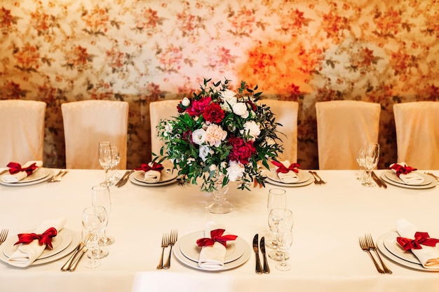 Una bella tavola servita con un bouquet chic sullo sfondo della parete tovagliolo bianco con fiocco rosso sul piatto e posate con bicchieri