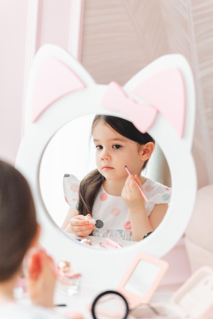Una bellissima bambina allo specchio che si pavoneggia una bambina è seduta a un tavolo per bambini e indossa cosmetici per bambini