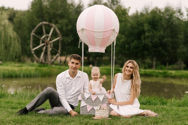 Una bellissima bambina con mamma e papà in un cestino con un palloncino e una torta festeggia il suo primo compleanno