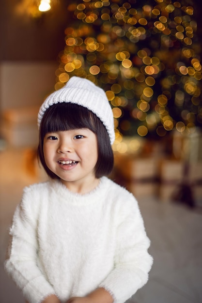 Una bellissima bambina coreana con un maglione bianco lavorato a maglia e un cappello in piedi sull'albero di Natale il giorno di Natale