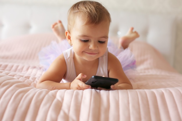 una bellissima bambina è sdraiata sul letto con una coperta rosa e tiene un telefono in mano