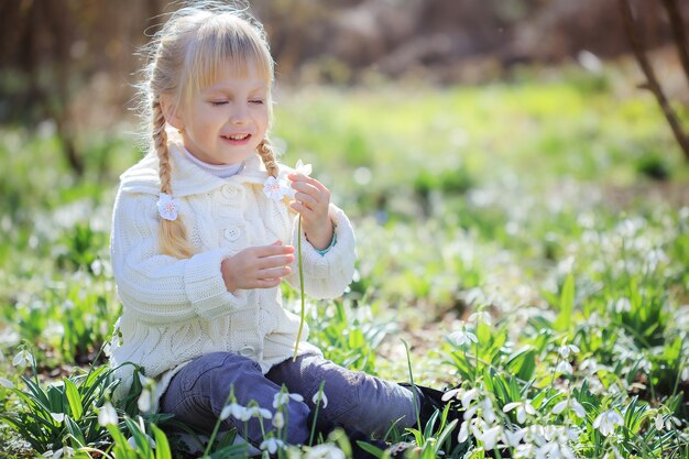 Una bellissima bambina è seduta su un prato fiorito. Una bambina con un maglione lavorato a maglia bianco sta prendendo in considerazione un bucaneve. Tempo di Pasqua. Foresta soleggiata di primavera