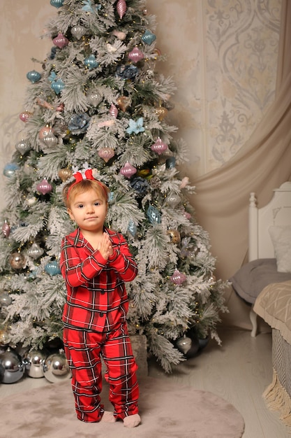 una bellissima bambina in pigiama natalizio rosso in una gabbia si trova all'albero di Natale