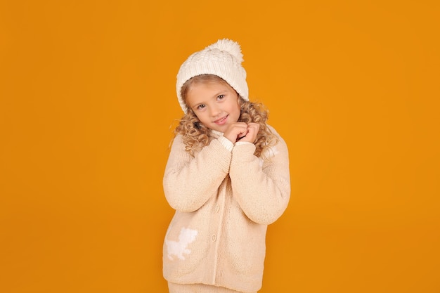 una bellissima bimba bionda con una giacca a maglia e un cappello a maglia invernale su uno sfondo giallo