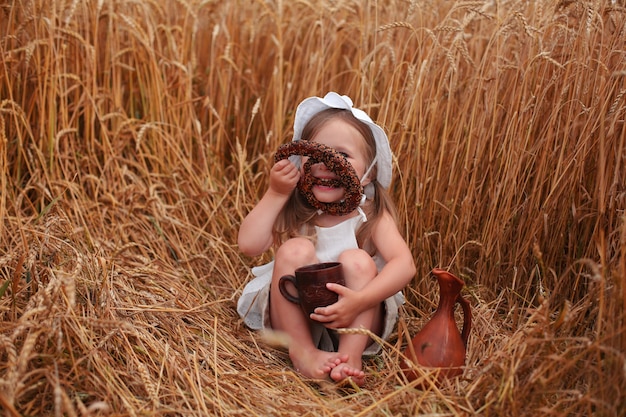 una bellissima bimba bionda è seduta in un campo di grano a mangiare pane e bere latte