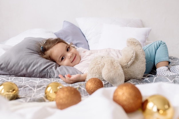 Una bellissima bimba di 2 anni dai capelli ricci è sdraiata sul letto tra luci e addobbi natalizi e ride