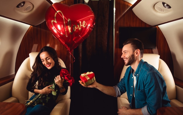 Una bellissima coppia sta festeggiando San Valentino su un jet privato. Un uomo sta facendo un regalo in una scatola rossa alla sua ragazza