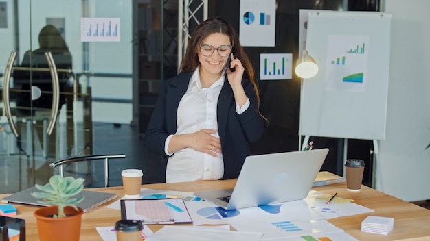 Una bellissima donna incinta che parla al cellulare seduta a un tavolo in un confortevole ufficio moderno