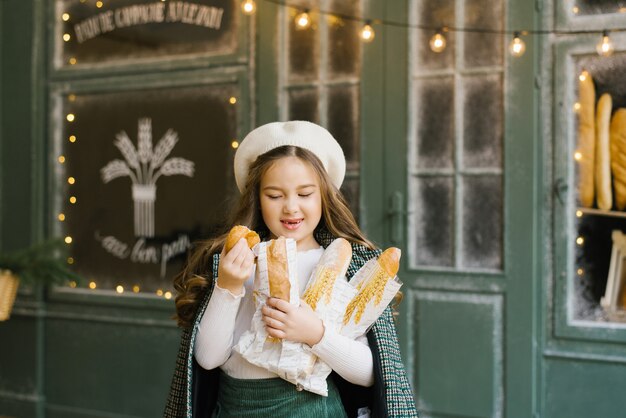 Una bellissima ed elegante bambina di sei anni tiene in mano una borsa di baguette e si trova vicino alla finestra di una panetteria con dolci freschi