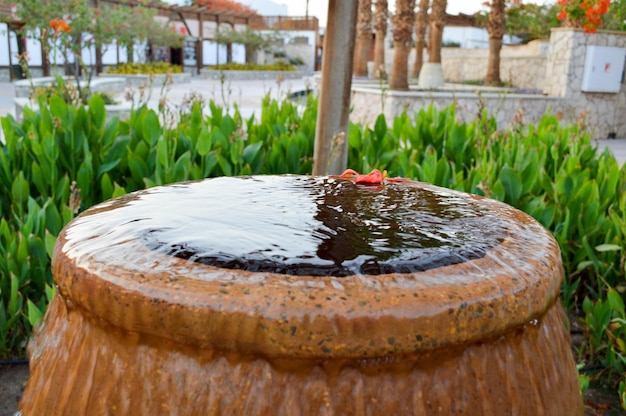 Una bellissima fontanella a forma di vaso marrone una brocca con gocce d'acqua che cadono su pietre colorate in piedi in un letto di fiori con piante verdi