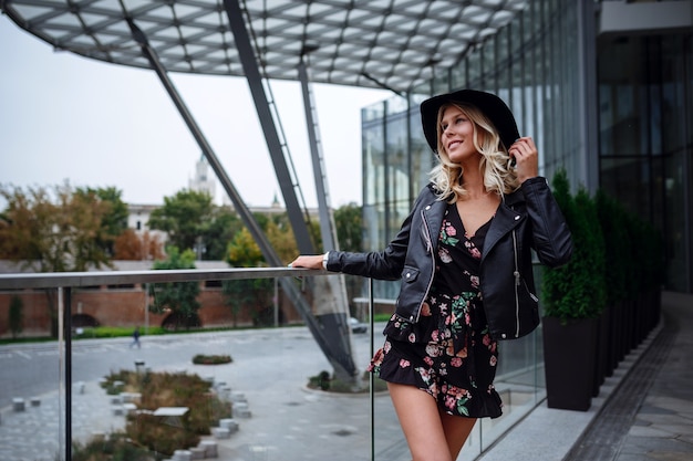 Una bellissima giovane donna bionda con una giacca di pelle nera, un bel vestito corto e un cappello cammina nel parco Zaryadye, Mosca.