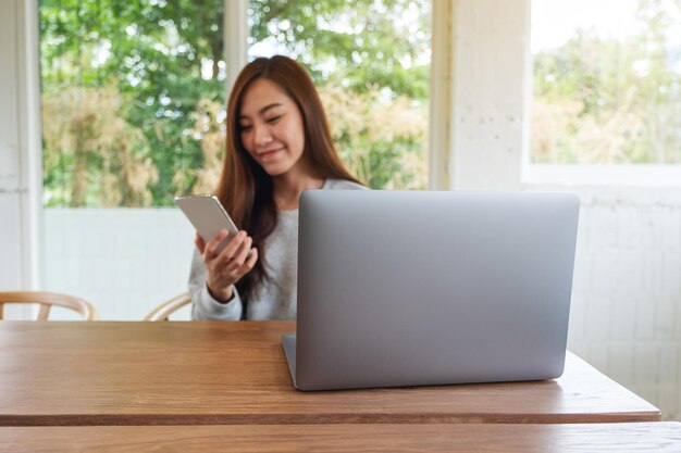 Una bellissima giovane donna che tiene in mano e usa il telefono cellulare con un computer portatile su un tavolo di legno