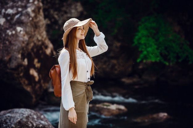 Una bellissima giovane donna con i capelli rossi, in un cappello e con uno zaino vicino a un fiume rumoroso nella foresta.