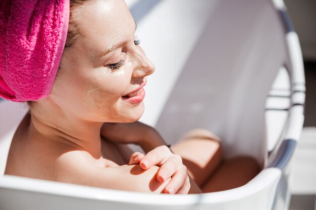 Una bellissima giovane donna con una maschera d'oro sul viso posa in una vasca da bagno in ceramica bianca