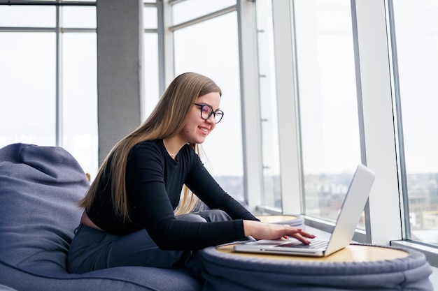 Una bellissima giovane manager siede con un laptop su un morbido pouf vicino alla finestra panoramica. Ragazza uomo d'affari che lavora a un nuovo progetto
