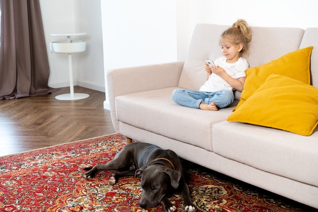 Una bimba bionda a casa sul divano con un cellulare e un grosso cane, bambini e gadget.