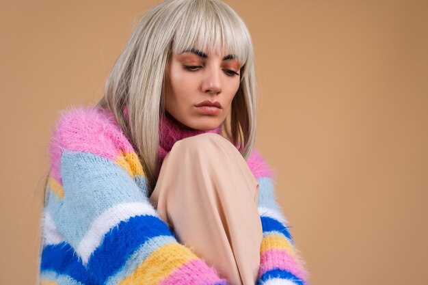 Una bionda con i capelli lunghi e la frangia, con il trucco nudo e in un soffice maglione multicolore luminoso su uno sfondo beige, la tristezza non sembra da nessuna parte