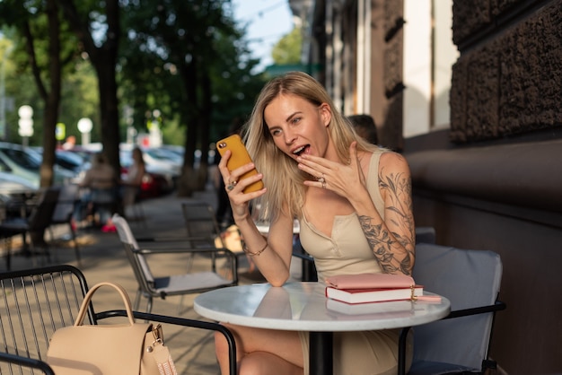 Una bionda con un tatuaggio sul braccio siede in un caffè estivo e parla al telefono con emozione