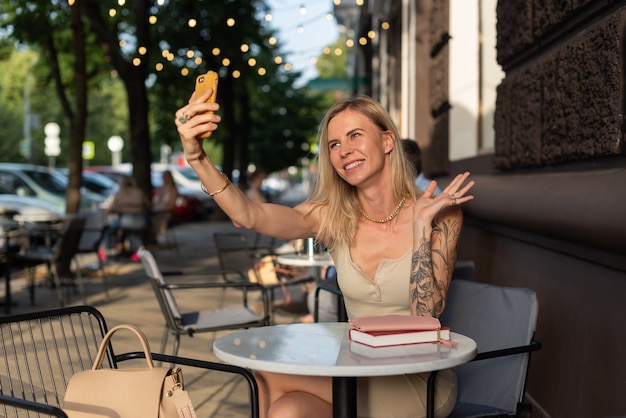 Una bionda con un tatuaggio sul braccio siede in un caffè estivo e parla al telefono con emozione