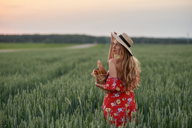 Una bionda con un vestito rosso e un cappello di paglia con del pane francese in mano su un campo di grano al tramonto