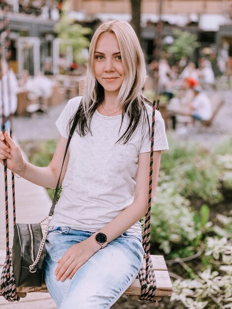 Una bionda europea con una maglietta bianca e jeans si siede su un'altalena sul territorio di una terrazza estiva di un caffè.