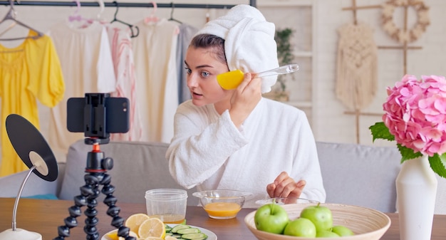 Una blogger di bellezza in una veste bianca con un asciugamano bianco sulla testa applica una maschera idratante verde su...