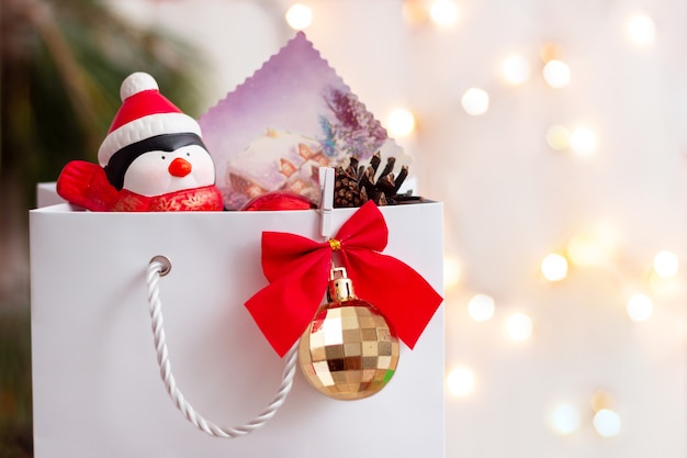 Una borsa di carta bianca con una palla d'oro un fiocco rosso con un pinguino giocattolo in un cappello di Babbo Natale e un biglietto di auguri