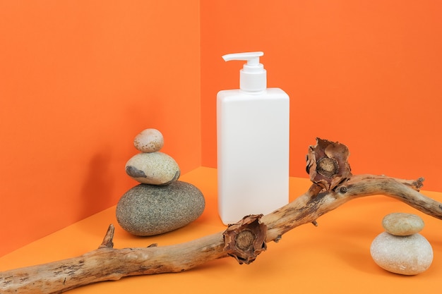 Una bottiglia cosmetica in bianco bianca con l'erogatore, rocce, bastone di legno con i fiori secchi nello spazio d'angolo sull'arancia
