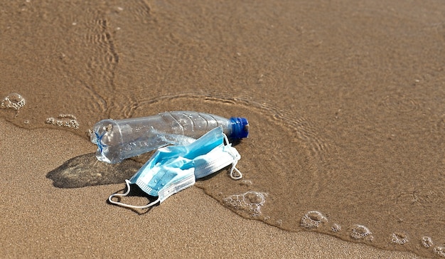 Una bottiglia di plastica giace e una maschera medica sulla spiaggia e inquina il mare