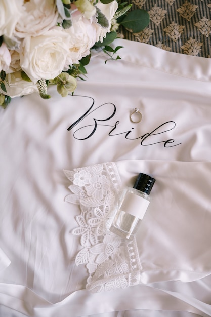 Una bottiglia di profumo con un tappo nero, una fede nuziale con una pietra e un bouquet da sposa di rose bianche