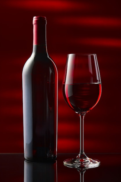 Una bottiglia e un bicchiere di vino rosso stanno su un tavolo a specchio nero. Sfondo nero e rosso.
