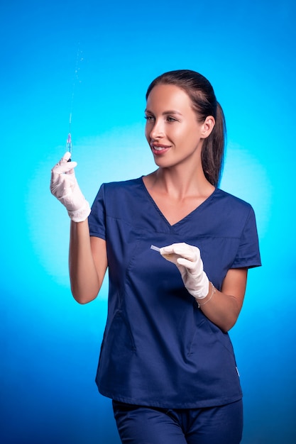 Una bruna con una coda intrecciata, in posa mentre si trovava su uno sfondo blu, in una tuta chirurgica blu in guanti medici, spremendo la medicina dalla siringa.