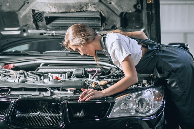 Una bruna con una tuta nera e una maglietta bianca vicino al cofano aperto dell'auto nera. Giovane donna in garage. concetto di riparazione auto