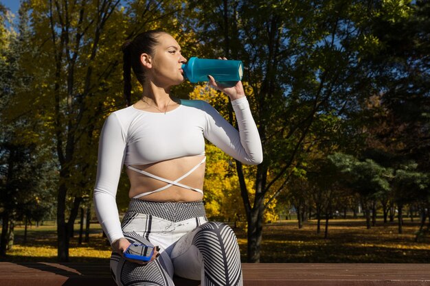 Una bruna in tuta beve acqua da una bottiglia dopo un allenamento nel parco Sport e stile di vita sano