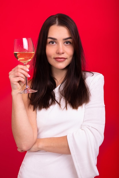 Una bruna in un abito bianco con un bicchiere di vino su fondo rosso Una giovane donna di buon umore celebra la vacanza